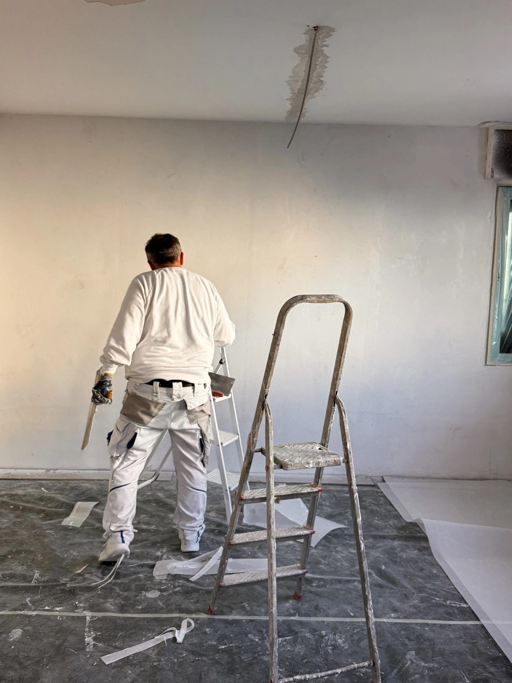 Handwerker bei Spachtelarbeiten an weißer Innenwand, Trittleiter und Schutzfolie auf Boden