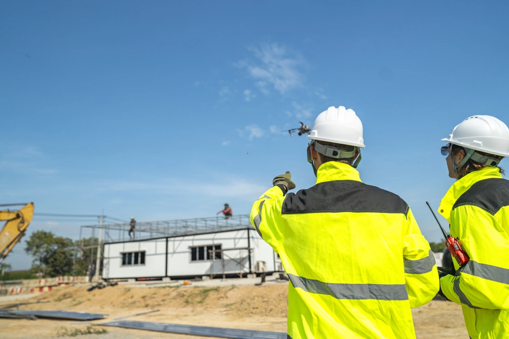 Zwei Bauleiter in Warnjacken prüfen Fassadenbau mit Drohne auf Baustelle